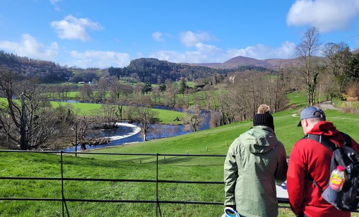 Two walkers with a dog approaching a filed overlooking a river and mountains